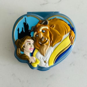 May include: A blue heart-shaped toy featuring Belle and the Beast from Beauty and the Beast. The toy has a blue background with a silhouette of a castle. Belle is wearing a yellow dress, and the Beast is brown with a white shirt.