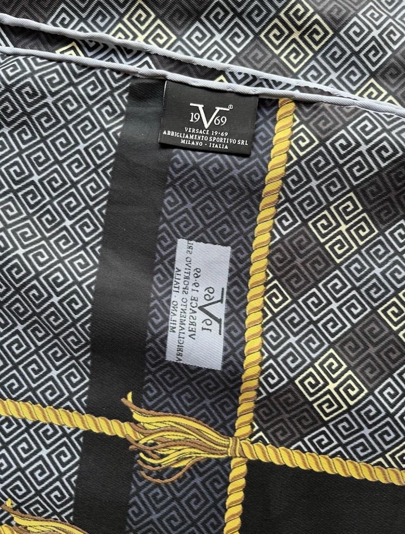 Peut inclure: Un foulard noir et blanc &agrave; motifs avec des accents de corde et de glands dor&eacute;s. Le foulard pr&eacute;sente un motif g&eacute;om&eacute;trique r&eacute;p&eacute;titif et la marque "Versace 19-69" est visible sur une &eacute;tiquette.