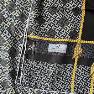 Peut inclure: Foulard &agrave; motifs noirs et dor&eacute;s avec un motif g&eacute;om&eacute;trique. Le foulard pr&eacute;sente des accents dor&eacute;s en forme de corde et des glands. L'&eacute;tiquette indique "Versace 1969". Le foulard a une bordure blanche.