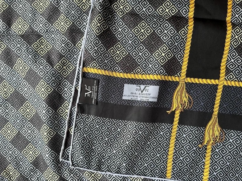 Peut inclure: Foulard &agrave; motifs noirs et dor&eacute;s avec un motif g&eacute;om&eacute;trique. Le foulard pr&eacute;sente des accents dor&eacute;s en forme de corde et des glands. L'&eacute;tiquette indique "Versace 1969". Le foulard a une bordure blanche.