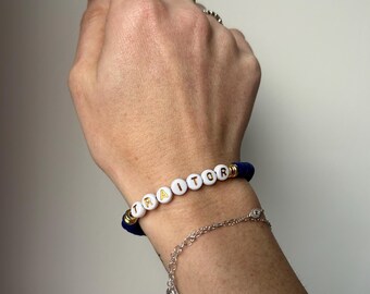 May include: A blue beaded bracelet with the word "TRAITOR" spelled out in white and gold letter beads. A silver chain bracelet is also visible. The bracelets are worn on a wrist.