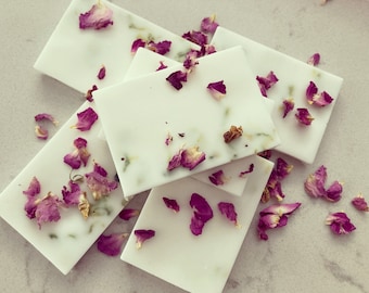 May include: A stack of white soap bars with dried rose petals and green herbs embedded in the soap. The soap bars are arranged on a white surface.