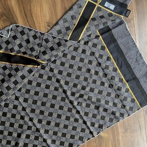 Peut inclure: &Eacute;charpe pli&eacute;e &agrave; motifs noirs et gris avec un motif g&eacute;om&eacute;trique. L'&eacute;charpe a une bordure noire avec une bordure en corde dor&eacute;e. L'&eacute;charpe est faite d'une mati&egrave;re douce et est pli&eacute;e sur une surface en bois.