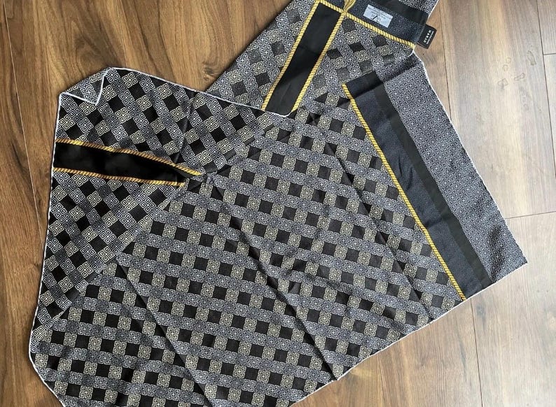 Peut inclure: &Eacute;charpe pli&eacute;e &agrave; motifs noirs et gris avec un motif g&eacute;om&eacute;trique. L'&eacute;charpe a une bordure noire avec une bordure en corde dor&eacute;e. L'&eacute;charpe est faite d'une mati&egrave;re douce et est pli&eacute;e sur une surface en bois.