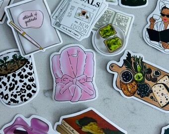 May include: A collection of stickers featuring various illustrations. Includes a pink robe, a notebook with "attitude of gratitude", a book with a Van Gogh painting, and a "GOALS" magazine. Other stickers depict food, coffee, and a hair clip.