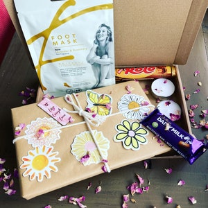May include: A brown gift box with a white string bow and a variety of colorful stickers. The box contains a foot mask, a chocolate bar, and a candle. The box is surrounded by purple flower petals.