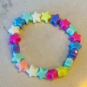 Può includere: Un braccialetto colorato a forma di stella con una chiusura. Il braccialetto è realizzato in colori pastello, tra cui rosa, blu, giallo e verde.