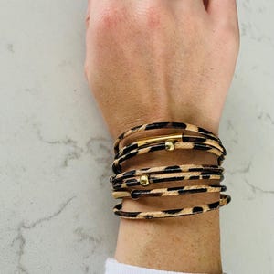 Puede incluir: Un conjunto de pulseras de varias capas con un estampado de leopardo y detalles dorados. Las pulseras están hechas de cuero y tienen pequeñas cuentas doradas y una barra dorada.