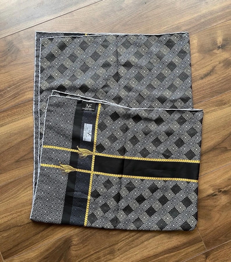 Peut inclure: Deux coussins d&eacute;coratifs noirs et gris &agrave; motifs g&eacute;om&eacute;triques. Un coussin a une bande noire avec une bordure en corde dor&eacute;e et des glands. Les coussins ont une bordure blanche et sont sur une surface en bois.