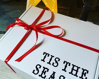 May include: A white gift box tied with a red ribbon. The words "TIS THE SEASON" are printed in black on the box. A star-shaped light is in the background.