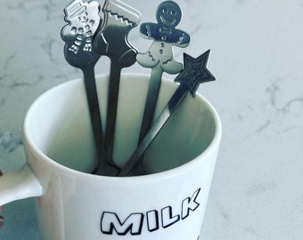 May include: A white ceramic mug with the words "MILK & COOKIES" in black lettering, holding four stainless steel Christmas-themed spoons. The spoons feature a snowman, stocking, gingerbread man, and star.