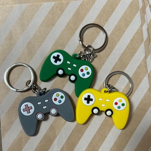 May include: Three colorful video game controller keychains. One is green, one is gray, and one is yellow. Each keychain has a silver ring and features a cross, buttons, and a joystick design.