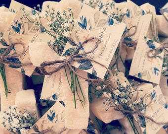 May include: A collection of small, white, rectangular soaps wrapped in twine and decorated with sprigs of white baby's breath flowers and blue and white floral patterned cards. The cards have the text "Chris's First Communion May 8th 2022".