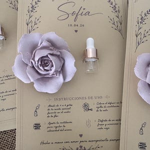 May include: A lavender-colored rose and a small glass bottle with a gold-colored ring are featured on a beige card. The card has text in Spanish, including "Sofia" and instructions for use. The overall theme is a wedding favor or gift.