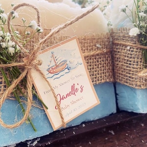 May include: Blue soap favors wrapped in burlap and tied with twine. Each favor has a small bouquet of white flowers and a tag that says "From My Shower To You, Janelle's Baby Shower, April 20, 2023."