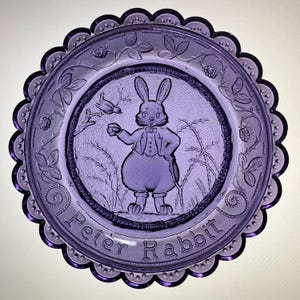 May include: A purple glass plate featuring a detailed embossed design. The center depicts Peter Rabbit in a suit, with the words "Peter Rabbit" below. The plate has a scalloped edge and is decorated with floral and berry motifs.