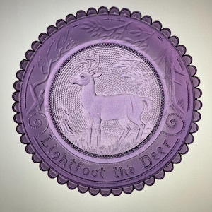 May include: A round purple glass plate with a scalloped edge featuring a central embossed image of a deer standing in a forest setting. The text Lightfoot the Deer is embossed along the bottom rim, surrounded by a decorative beaded border.