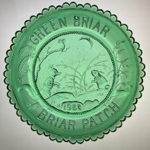 May include: Green glass plate with a scalloped edge. The plate features an embossed design of an owl, fox, and rabbit in a briar patch setting, with the words "GREEN BRIAR" and "BRIAR PATCH" and the year "1982".