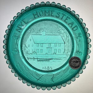May include: A teal-colored glass plate with a scalloped edge. The plate features an embossed image of a house with the text "NYE HOMESTEAD" and the year "1685". A Fairpoint Glass Company sticker is visible.
