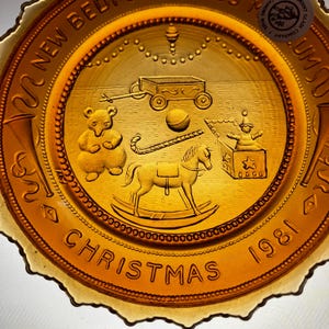 May include: A vintage amber glass Christmas plate with a raised design of toys including a rocking horse, a toy wagon, a teddy bear, a ball, and a toy box with a doll inside. The plate has a scalloped edge and the words "2000 NEW BEGINNINGS CHRISTMAS 1981" around the rim.