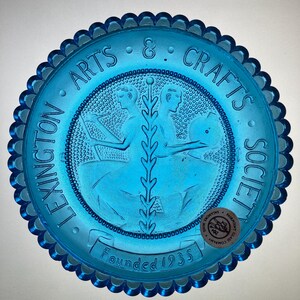 May include: A round, translucent blue glass plate with a scalloped edge. The center features embossed figures of two people and a central plant motif. The text reads LEXINGTON ARTS & CRAFTS SOCIETY and Founded 1935. A circular sticker is attached.