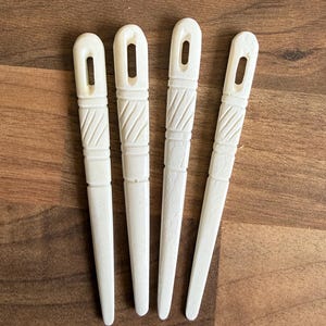 Könnte beinhalten: Vier cremefarbene Nadeleinfädler aus knochenähnlichem Material liegen auf einer Holzoberfläche. Jeder hat ein rechteckiges Loch oben und ein geschnitztes Muster am Griff. Die Einfädler sind konisch und scheinen Vintage zu sein.