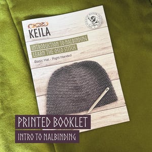 以下が含まれることがあります： Introduction to Nalbinding Learn the Oslo Stitch Basic Hat - Right Handedというタイトルの印刷された説明用小冊子。表紙には、明るい木目調の背景に、ダークグレーの質感のある帽子と針の写真が掲載されており、Keilaのロゴも含まれています。