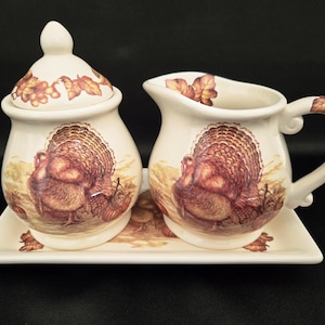 May include: Cream-colored ceramic sugar bowl with lid and creamer set on a rectangular tray. Each piece features a detailed illustration of a turkey and autumn foliage. The set is designed with a Thanksgiving theme.