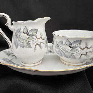 May include: A white porcelain tea set with a creamer, sugar bowl, and serving tray. The set features a gray and beige leaf design with gold trim. The creamer has a curved handle and spout.