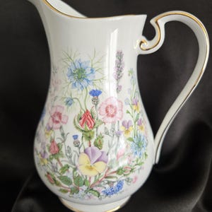 May include: White ceramic pitcher with a floral design featuring blue, pink, purple, and yellow flowers. The pitcher has a gold trim on the handle and spout. The pitcher is a decorative item.
