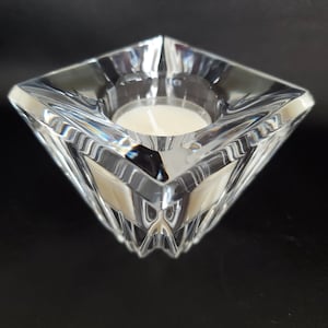 May include: A clear glass candle holder with a square base and a faceted design. The holder is designed to hold a small, round candle.