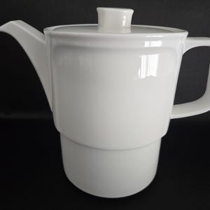 May include: A white ceramic teapot with a lid, spout, and handle. The teapot has a cylindrical base and a slightly curved body. The handle is a simple loop design. The teapot is approximately 8 inches tall.