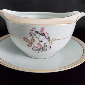 May include: A white porcelain gravy boat with a gold trim and a floral design in shades of pink, gray, and gold. The boat is sitting on a matching saucer.