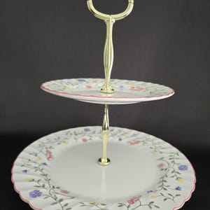 May include: Two-tiered serving stand with a gold-colored handle. The white plates feature a floral pattern with pink, blue, and yellow flowers. The edges of the plates have a pink scalloped trim. Perfect for displaying desserts or appetizers.