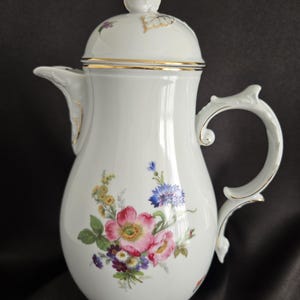 May include: White porcelain coffee pot with a lid, featuring a floral design in pink, blue, yellow, and purple. The pot has a gold trim and handle. The lid is topped with a small, white, flower-shaped knob.
