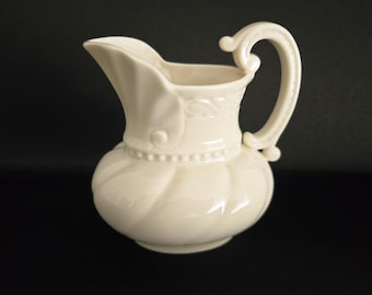 May include: Cream-colored ceramic pitcher with a bulbous body and a curved handle. The pitcher features decorative embossed details around the top and base, and a spout for pouring. The handle has a decorative swirl at the top.