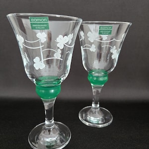 May include: Two clear glass goblets with a green base and stem, adorned with white clover designs. The goblets have a flared rim and a label that reads "eamon HAND ENGRAVED IN IRELAND".