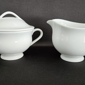 May include: A white ceramic sugar bowl with a lid and a creamer. The sugar bowl has two handles and a loop handle on the lid. The creamer has a handle and a spout. Both pieces have a pedestal base.