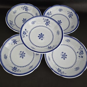 May include: Five white ceramic bowls with a blue floral design. Each bowl has a scalloped edge and a blue border. The floral pattern includes a central flower and smaller floral clusters around the rim. The bowls are arranged on a dark surface.
