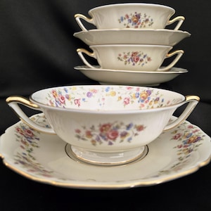 May include: Cream-colored porcelain soup bowls and saucers with floral patterns. The bowls have two handles with gold trim, and the saucers have scalloped edges. The floral design includes pink, blue, and yellow flowers.