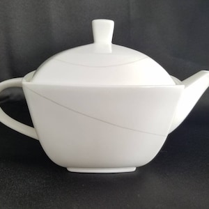 May include: White porcelain teapot with a lid and a curved spout. The teapot has a modern design with a subtle gray line detail.