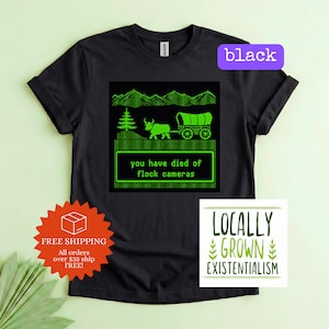May include: A black short-sleeved t-shirt featuring a bright green pixelated graphic of a covered wagon, a tree, and a mountain range. The text within the graphic reads, you have died of flock cameras. The shirt has a crew neckline and short sleeves.