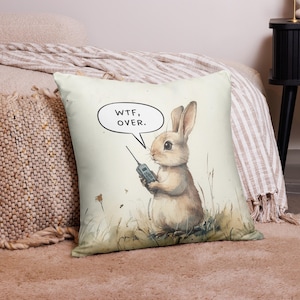 May include: Square decorative pillow featuring a watercolor illustration of a rabbit holding a walkie-talkie. The rabbit is in a grassy field, with the text "WTF, OVER." in a speech bubble. The pillow has a neutral background.