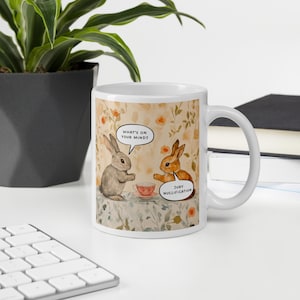 Puede incluir: Taza de cerámica blanca con una ilustración humorística de dos conejos. Uno pregunta "WHAT'S ON YOUR MIND?" y el otro responde "JURY NULLIFICATION". La taza tiene un fondo floral y una taza de té.