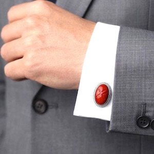 Peut inclure: Un bouton de manchette ovale en argent avec une incrustation de pierre rouge profonde et lisse ornée d'une croix gravée. Le bouton de manchette est fixé sur une manchette de chemise blanche, partiellement recouverte par une veste de costume grise.