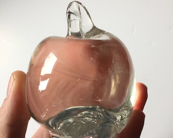 May include: A clear glass apple with a stem. The apple is smooth and has a slight pink hue.