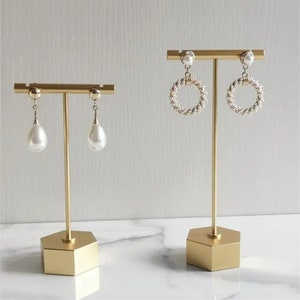May include: Two gold metal earring stands with hexagonal bases. One stand displays a pair of pearl drop earrings, while the other stand displays a pair of pearl hoop earrings.