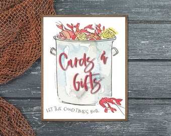 May include: A white sign with a watercolor illustration of a pot of crawfish and the text "Cards & Gifts" and the phrase "Let the good times boil".