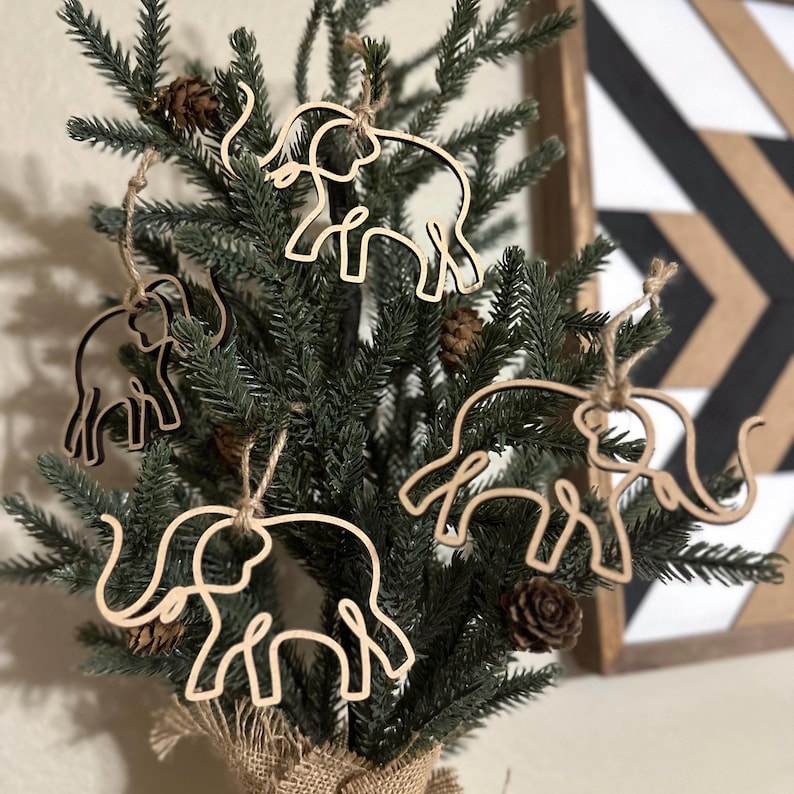 Puede incluir: Un peque&ntilde;o &aacute;rbol de Navidad decorado con adornos de elefantes de madera. Los adornos son de color marr&oacute;n claro y tienen un dise&ntilde;o sencillo de l&iacute;nea continua. El &aacute;rbol est&aacute; adornado con pi&ntilde;as y se encuentra en una maceta envuelta en arpillera.
