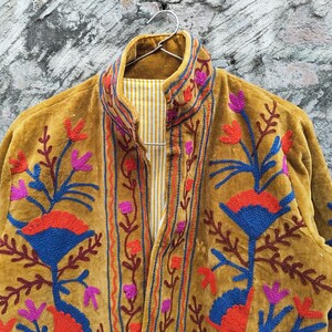 May include: Mustard-coloured jacket with detailed floral embroidery in red, blue, and purple. The jacket has a high collar and a striped lining. The embroidery provides a striking contrast against the jacket's textured fabric, showcasing a bohemian style.